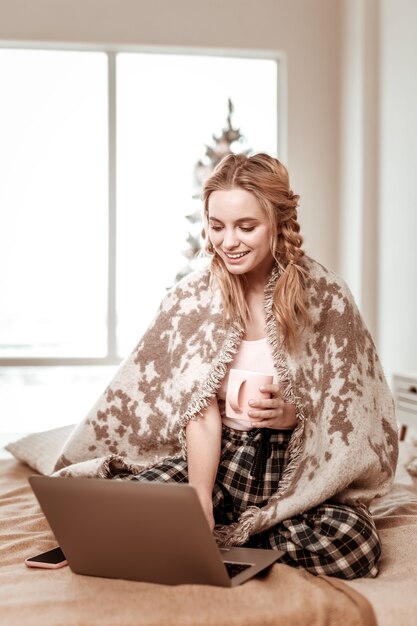 Pokryty kratą. Zadowolona dziewczyna ze skomplikowaną fryzurą siedzi na łóżku w piżamie i sprawdza laptopa