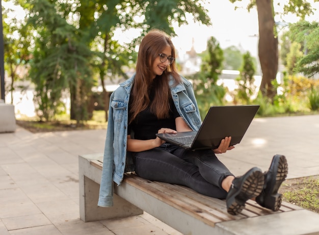 Połączenie online. Nauka na odległość. Młoda kobieta studentka w dżinsowej kurtce i okularach patrzy na ekran laptopa siedząc na ławce w parku