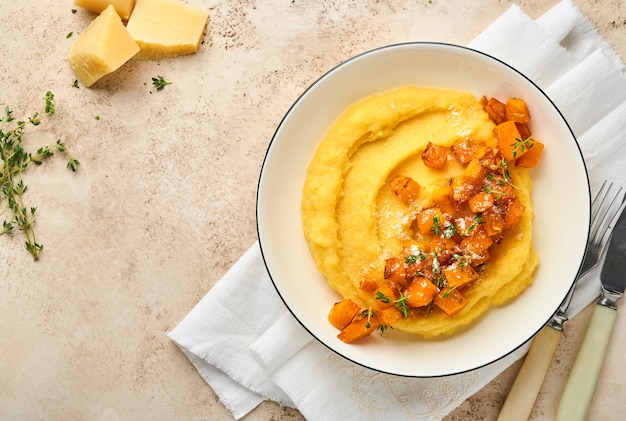 Polenta z masłem, dynią, czosnkiem, cząberem i parmezanem w białej misce na jasnym betonowym tle. Tradycyjne podstawowe włoskie jedzenie, jedzenie wegańskie. Widok z góry. Skopiuj miejsce.