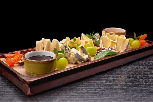 Półmisek serów na drewnianej desce z winogronami, miętą, miodem i dżemem, na czarnym stole, widok z boku