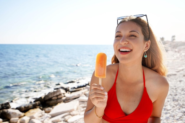 Południowoamerykańska śmiejąca się dziewczyna jedząca pomarańczowe lody na patyku na plaży latem