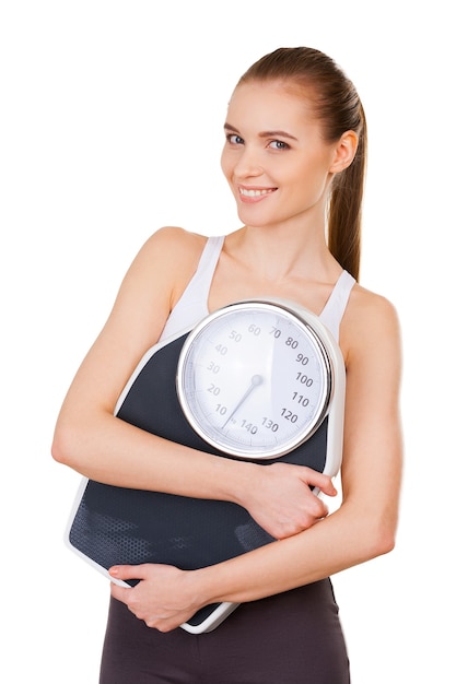 Pomaga mi kontrolować wagę. Atrakcyjna młoda kobieta w odzieży sportowej trzyma wagę i uśmiecha się stojąc na białym tle