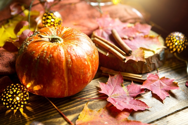 Pomarańczowa naturalna okrągła dynia na drewnianym stole z opadłymi żółtymi i czerwonymi liśćmi klonu, laski cynamonu. Girlandy świetlne, ciepła jesienna atmosfera, Święto Dziękczynienia, dożynki, Halloween. Skopiuj miejsce