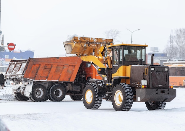 Pomarańczowy ciągnik czyści śnieg z drogi i ładuje go do ciężarówki. Sprzątanie dróg
