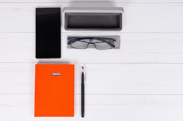 Pomarańczowy pamiętnik z długopisem do organizowania harmonogramu, okulary i otwarte etui na okulary przy telefonie komórkowym na drewnianym białym stole