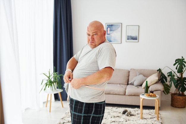 Pomiar talii Śmieszny mężczyzna z nadwagą w codziennych ubraniach jest w domu w domu