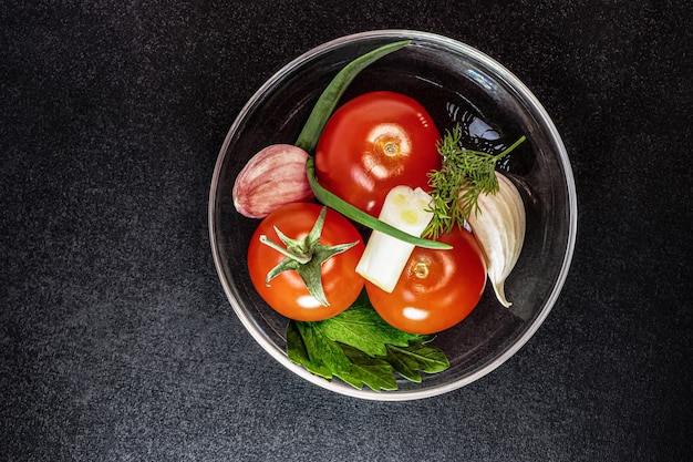 Pomidory czereśniowe z czosnkiem i cebulą i młodymi ziołami na szklanej płytce. Czarne tło.