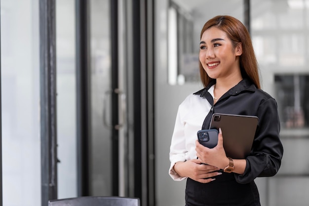 Pomyślna bizneswoman trzyma tabletkę i telefon komórkowy stojący w biurze