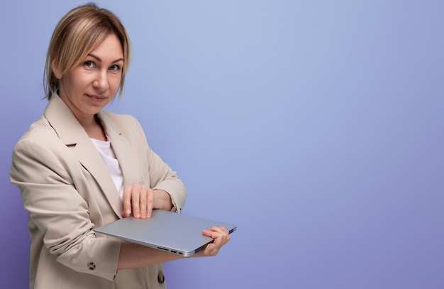 Pomyślna blond młoda kobieta w biznesowej kurtce z laptopem na pracownianym tle
