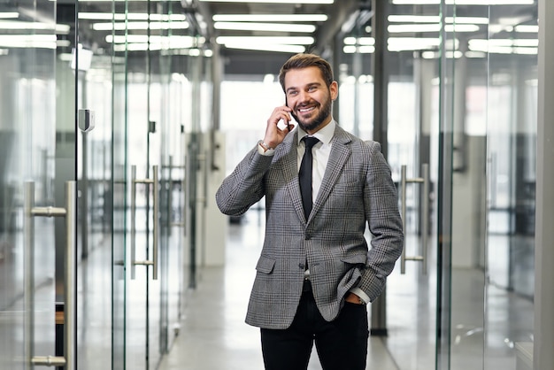 Pomyślny biznesowy mężczyzna ma rozmowę telefoniczną podczas gdy stojący w pustym biurowym korytarzu. Męski profesjonalny bankowiec opowiada na telefonie komórkowym podczas pracy przerwy w kostiumu.