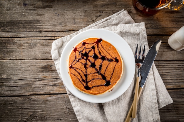 Pomysły na śniadanie to Halloween, jedzenie dla dzieci. Placki z dyni udekorowane syropem czekoladowym w tradycyjnym stylu - pajęczyna, pająk, latarnia jack. Na drewnianym stole w stylu rustykalnym, miejsce