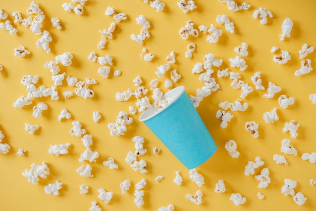 Popcorn w niebieskim papierowym kubku rozrzuconym na żółtym tle. Minimalistyczna koncepcja. Widok z góry. Kopiuj, puste miejsce na tekst.