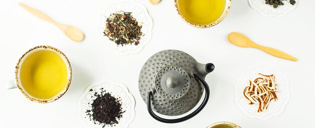 Popielaty żelazny czajnik wśród różnych rodzajów sucha herbata i filiżanki z gotowym napojem na białym stole