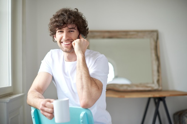 Poranna kawa w domu Ujęcie szczęśliwego młodego mężczyzny rozmawiającego przez telefon komórkowy i pijącego filiżankę kawy siedzącego w salonie w domu