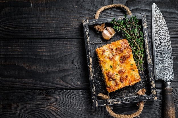 Porcja lasagne z mielonym mięsem wołowym i pomidorowym sosem bolońskim na drewnianej tacy. Czarne drewniane tło. Widok z góry. Skopiuj miejsce.