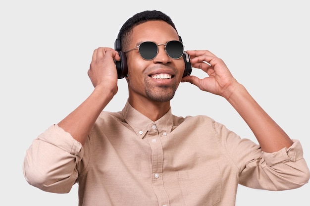 Portret African American Młody Człowiek Ze Słuchawkami Słuchać Muzyki I Tańca Szczęśliwy Uśmiechający Się Szeroko Afro Mężczyzna W Lustrzanych Okularach Przeciwsłonecznych I Swobodnym Stroju Pozowanie Na Tle Białego Studia
