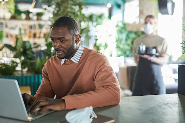 Portret afroamerykańskiego biznesmena korzystającego z laptopa podczas pracy w ekologicznym wnętrzu kawiarni ozdobionym świeżymi zielonymi roślinami, kopia przestrzeń