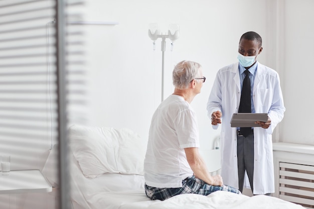 Portret afroamerykańskiego lekarza rozmawiającego ze starszym mężczyzną w białej sali szpitalnej, kopia przestrzeń