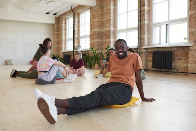 Portret afrykańskiego mężczyzny uśmiecha się do kamery siedzi na podłodze w studio tańca z innymi ludźmi