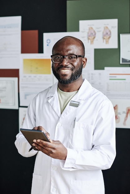 Portret afrykańskiego, odnoszącego sukcesy lekarza w białym fartuchu, uśmiechającego się do kamery podczas korzystania z komputera typu tablet podczas w