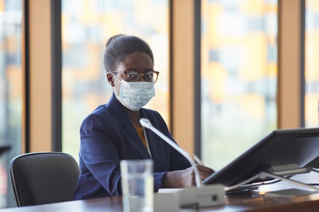 Portret afrykańskiej bizneswoman w okularach i masce ochronnej patrząc, siedząc przy stole w sali konferencyjnej