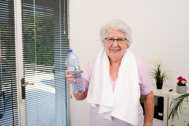 Portret aktywnej i dynamicznej starszej kobiety uprawiającej sport fitness w domu i trzymającej butelkę wody mineralnej