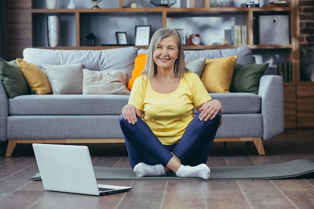 Portret aktywnej starszej kobiety siedzącej w domu na podłodze lotosu i patrzącej na kamerę szczęśliwa babcia robi jogę online za pomocą laptopa