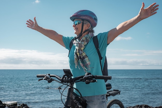 Portret aktywnej starszej kobiety z wyciągniętymi ramionami, ciesząc się słonecznym dniem na plaży z rowerem elektrycznym Autentyczna koncepcja życia na emeryturze i zdrowego stylu życia Horyzont nad morzem