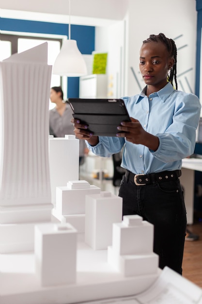 Portret architekta pracującego na tablecie stojącym obok architektonicznej makiety kompleksu budowlanego budynków biurowych. Inżynier porównujący plany na urządzeniu cyfrowym z modelem piankowym w skali.