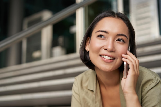 Portret asian uśmiecha się dziewczyna podczas rozmów na telefon komórkowy Młoda kobieta dzwoni do znajomego siedzącego na schodach