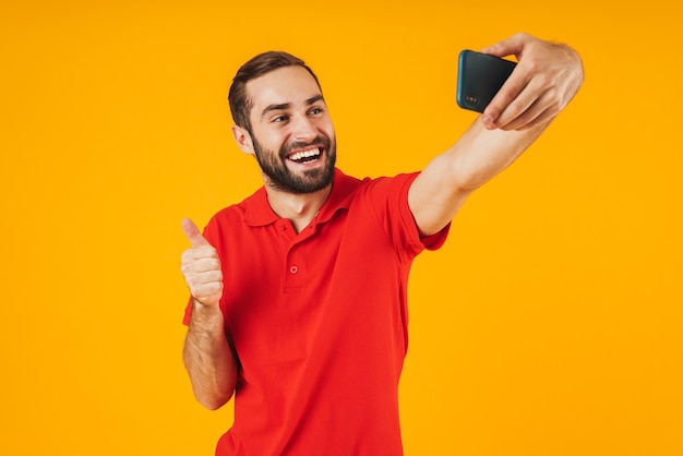 Portret atrakcyjnego mężczyzny w czerwonej koszulce śmiejącego się i pokazującego kciuk podczas robienia zdjęcia selfie na telefonie komórkowym na białym tle nad żółtym