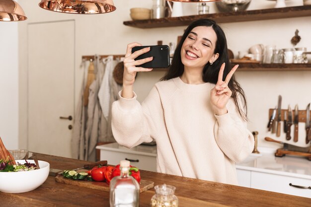 Portret atrakcyjnej brunetki kobiety robienie zdjęć selfie na smartfonie podczas gotowania zielonej sałatki z warzywami w stylowej kuchni w domu