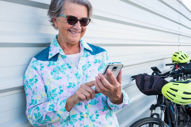 Portret atrakcyjnej i uśmiechniętej starszej kobiety stojącej przed białą metalową ścianą i wiadomościami z jej telefonu komórkowego. Dwa rowery elektryczne w kolorze czarnym blisko niej