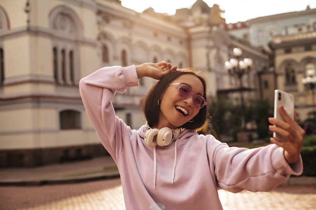 Portret atrakcyjnej młodej kobiety w stylowych różowych słuchawkach z kapturem i okularach przeciwsłonecznych uśmiecha się i robi selfie na zewnątrz