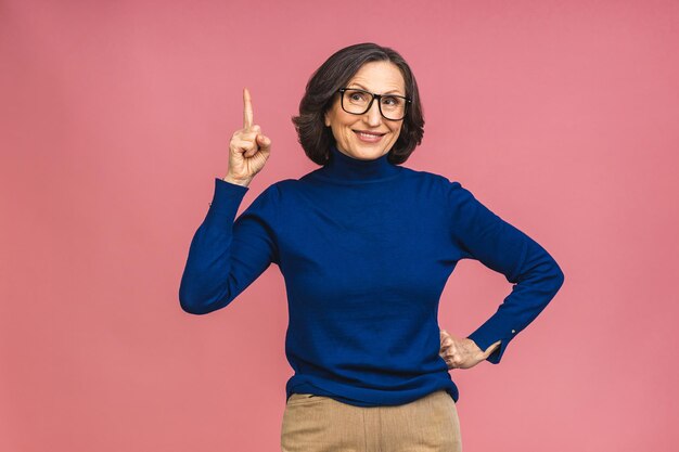 Portret atrakcyjnej wesołej pięknej starszej dojrzałej kobiety w wieku demonstrującej miejsce na kopię na białym tle nad różowym tłem, wskazując palcem w górę.