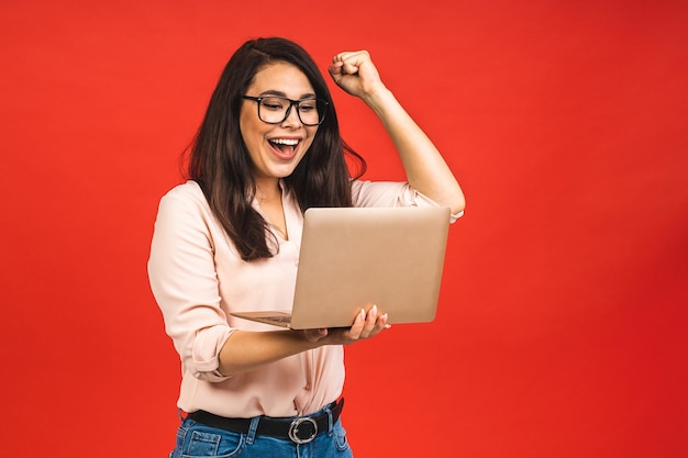 Portret atrakcyjnej zdziwionej podekscytowanej uśmiechniętej kobiety biznesu trzyma rękę patrząc na ekran laptopa na białym tle na czerwonym tle Zwycięzca bizneswoman z sukcesem