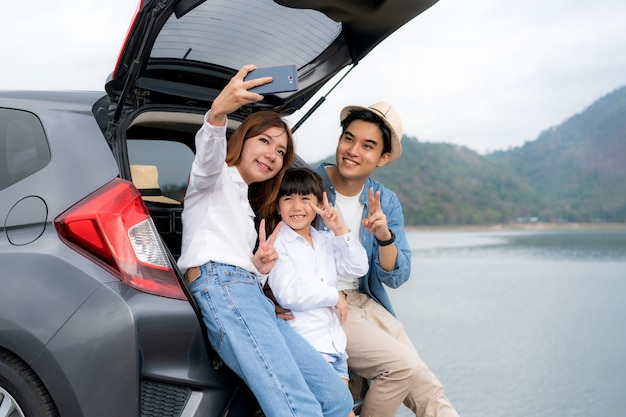 Portret Azjatycki rodzinny obsiadanie w samochodzie z ojca, matki i córki selfie z widokiem jeziora i góry smartphone, podczas gdy wakacje wpólnie w wakacje. Szczęśliwego rodzinnego czasu.