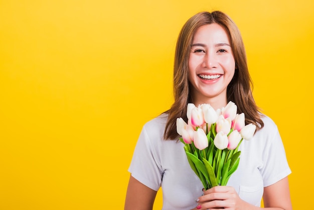 Portret azjatycki tajski piękna szczęśliwa młoda kobieta uśmiechając się, krzycząc podekscytowany trzymać kwiaty tulipany bukiet w ręce i patrząc do kamery, studio strzał na białym tle na żółtym tle, z miejsca na kopię