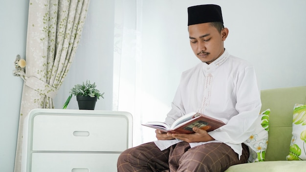 Portret azjatyckiego muzułmanina siedzącego na kanapie i czytającego Koran w domu