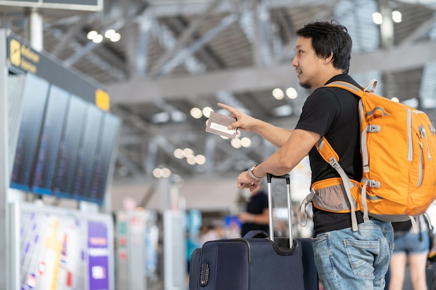 Portret azjatyckiego podróżnika z bagażem, z paszportem patrzącym i wskazującym na tablicę lotów