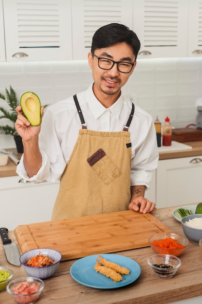 Portret azjatyckiego uśmiechniętego szefa kuchni w mundurze krojenia awokado do robienia sushi