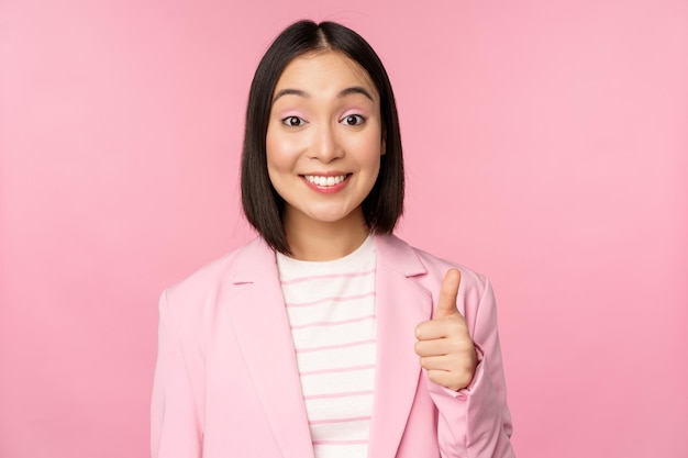 Portret azjatyckiej bizneswoman uśmiechniętej zadowolonej pokazującej kciuki do góry chwalące jak i aprobujące stojące w garniturze na różowym tle