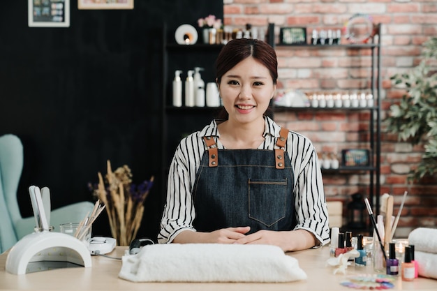 Portret azjatyckiej koreańskiej manikiurzystki w salonie spa. sukcesy pewnie salon kosmetyczny właściciele kobiet patrząc na kamery siedząc w studio. Wesoła kosmetyczka uśmiecha się i czeka na klienta w warsztacie.