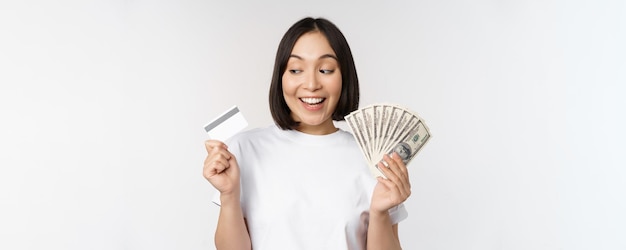 Portret azjatykciej kobiety uśmiechający się trzymający kartę kredytową i pieniądze gotówkowe dolary stojące w tshirt nad białym tłem