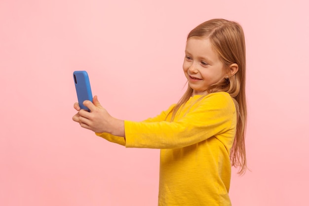 Portret Bardzo Pięknej Małej Dziewczynki Z Piegami I Rudymi Włosami Uśmiecha Się Do Smartfona, Biorąc Selfie Lub Komunikując Się Z Rodzicami Podczas Rozmowy Wideo W Studio Strzał Na Białym Tle Na Różowym Tle