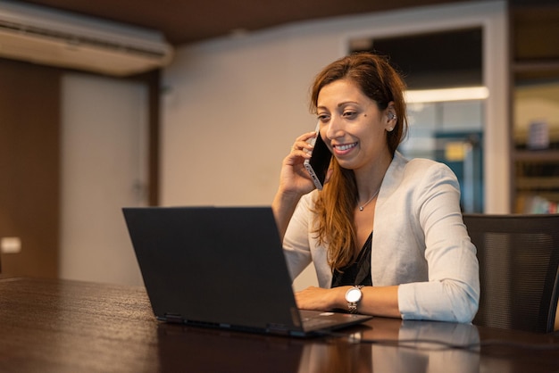 Portret bizneswoman używającej laptopa w nocy w biurze podczas rozmowy przez telefon