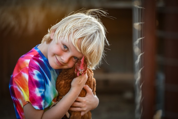 Portret blond chłopiec rasy kaukaskiej w kolorowej koszuli, przytulający uroczą kurę