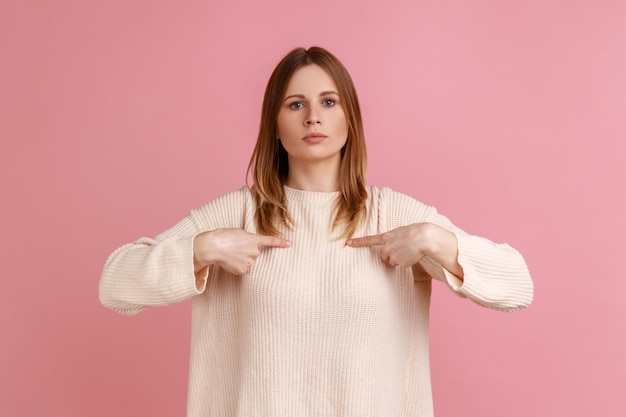 Zdjęcie portret blond wyniosły kobiety, wskazując na siebie i patrząc z arogancją na aparat, będąc dumnym i samolubnym, nosząc biały sweter kryty studio strzał na białym tle na różowym tle