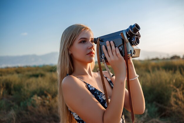 Portret blondynki kobieta w kwiecistej druk sukni z rocznik kamerą wideo w winogrona polu podczas zmierzchu