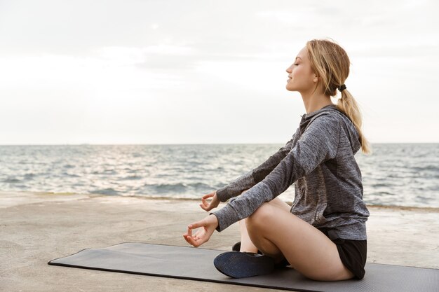 Portret blondynki zrelaksowanej kobiety noszącej dres siedzący z gestem zen podczas medytacji na macie do jogi w pobliżu morza rano